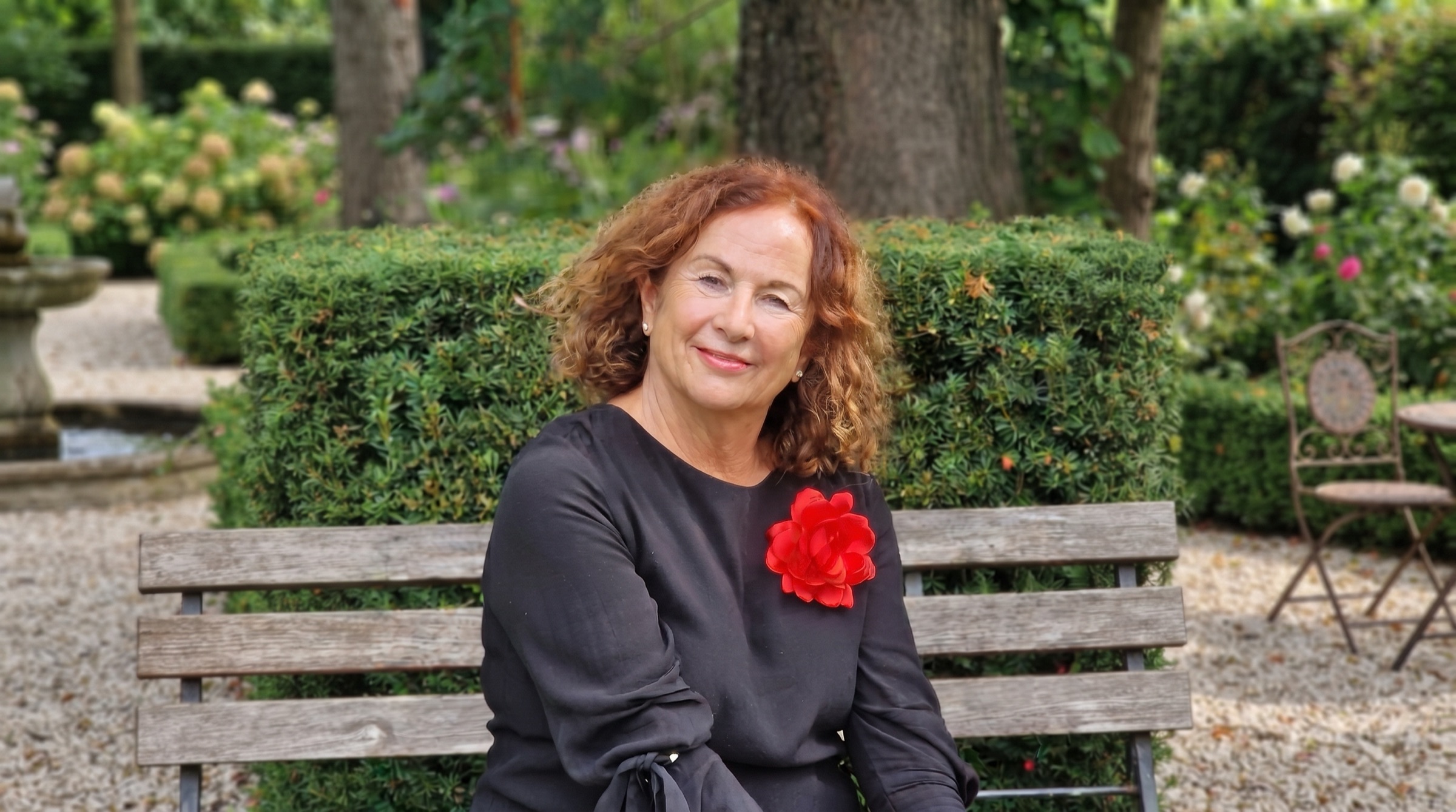 Lisa Kircher sitzt auf einer Holzbank in einem Garten, hinter ihr eine Hecke, an ihrem Kleid eine rote Blüte