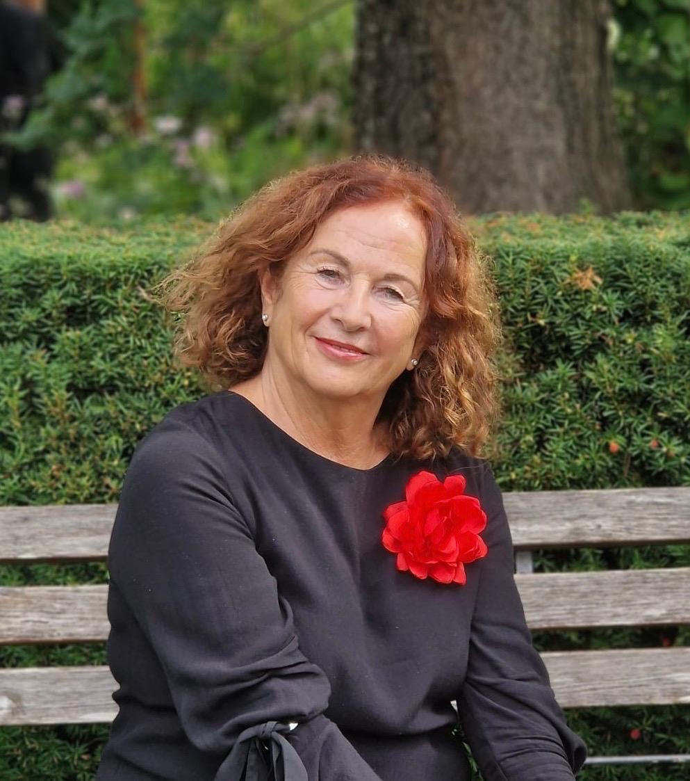 Lisa Kircher sitzt auf einer Holzbank vor einer Hecke, mit einer roten Blüte am Kleid
